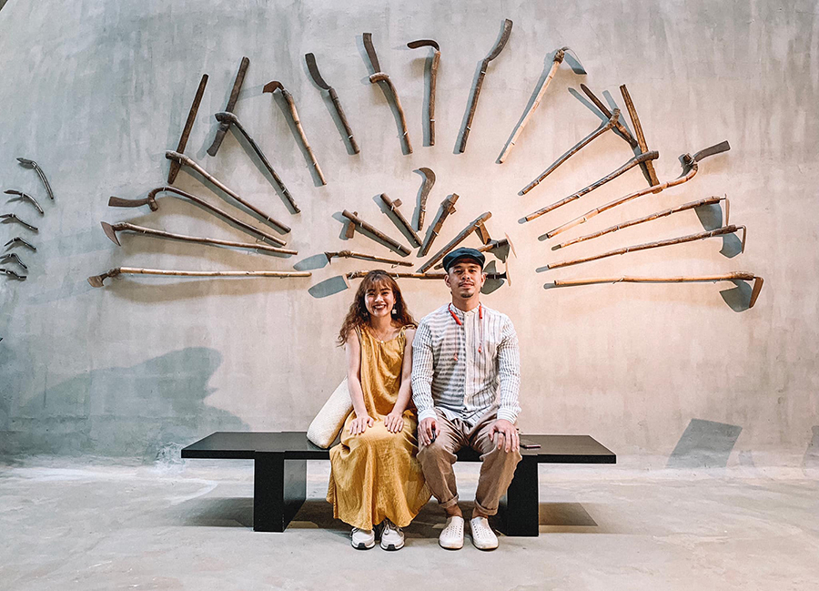 Hai người ngồi tr&ecirc;n ghế trước bức tường trang tr&iacute; bằng nhiều c&ocirc;ng cụ n&ocirc;ng nghiệp truyền thống.