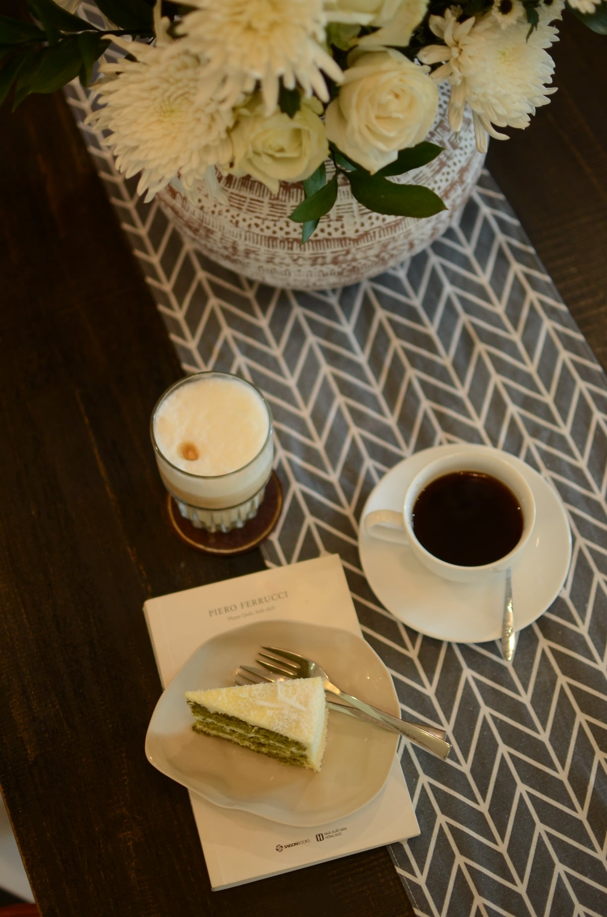 Ly c&agrave; ph&ecirc;, ly sữa, đĩa b&aacute;nh v&agrave; quyển s&aacute;ch tr&ecirc;n b&agrave;n c&oacute; khăn trải hoạ tiết, k&egrave;m b&igrave;nh hoa c&uacute;c trắng.