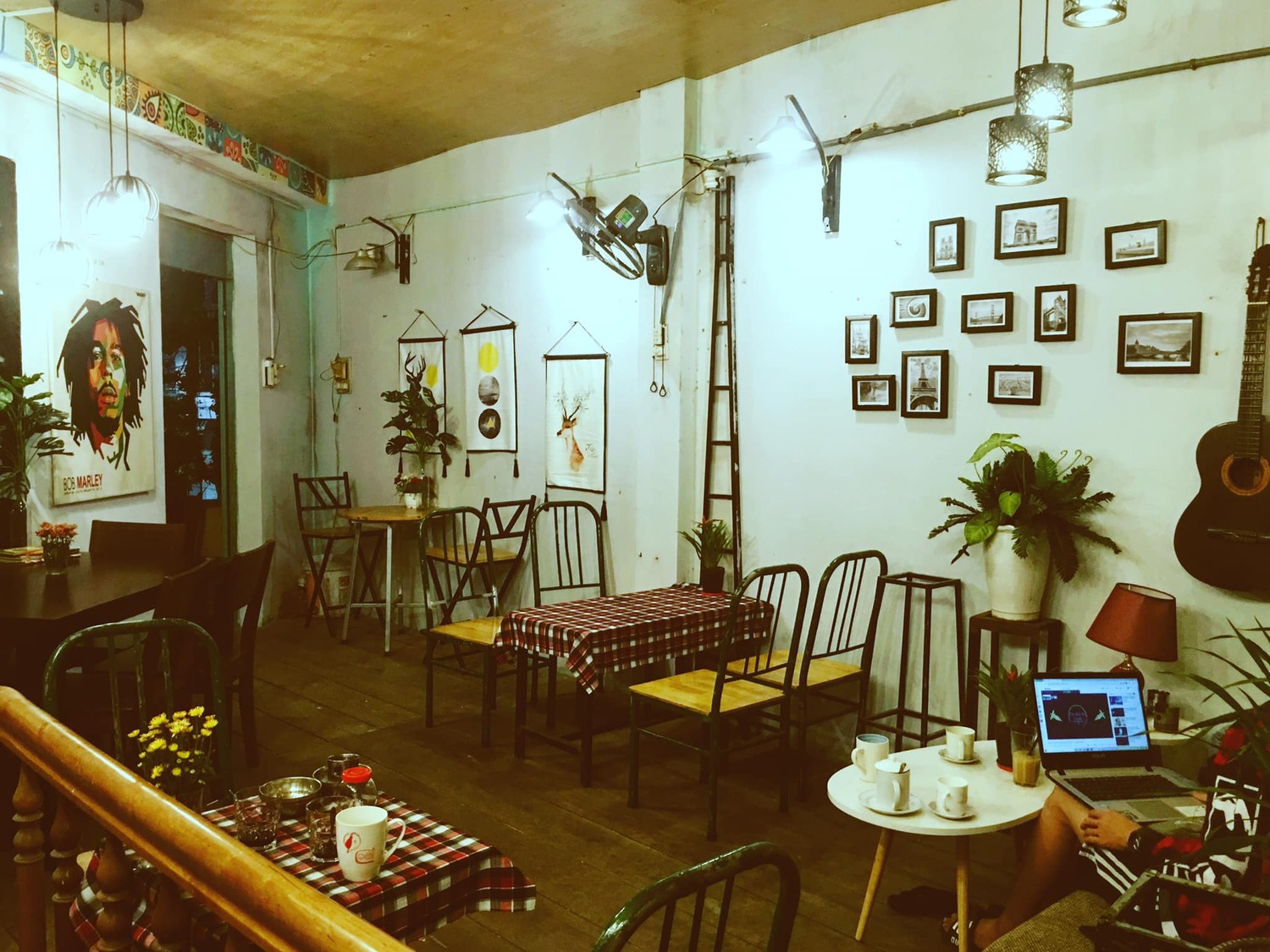 Qu&aacute;n c&agrave; ph&ecirc; ấm c&uacute;ng với b&agrave;n ghế gỗ, tranh treo tường, đ&agrave;n guitar, &aacute;nh s&aacute;ng dịu nhẹ, v&agrave; người đang sử dụng laptop.