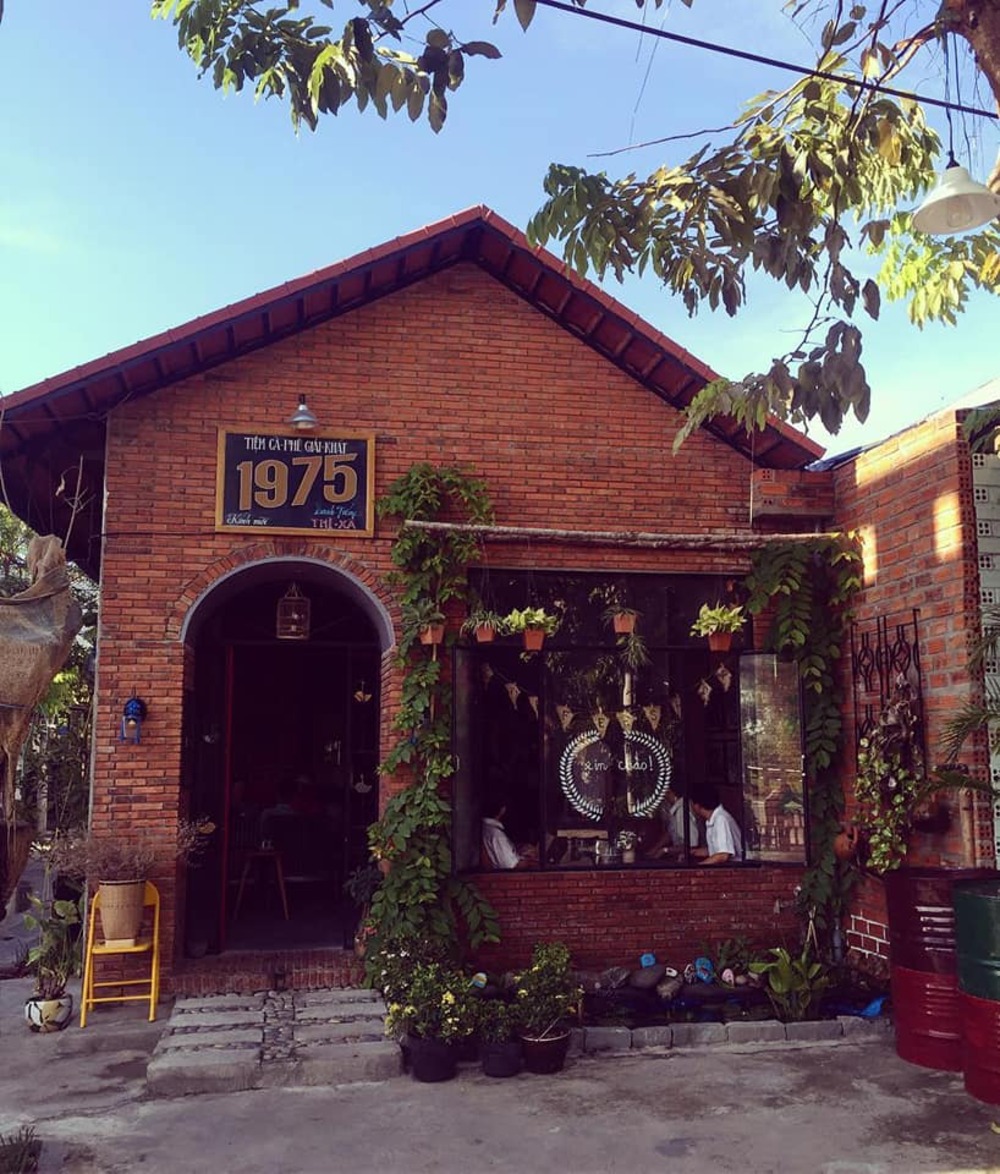 Qu&aacute;n c&agrave; ph&ecirc; với tường gạch đỏ, cửa sổ k&iacute;nh lớn v&agrave; c&acirc;y xanh trang tr&iacute;, biển hiệu ghi "1975".