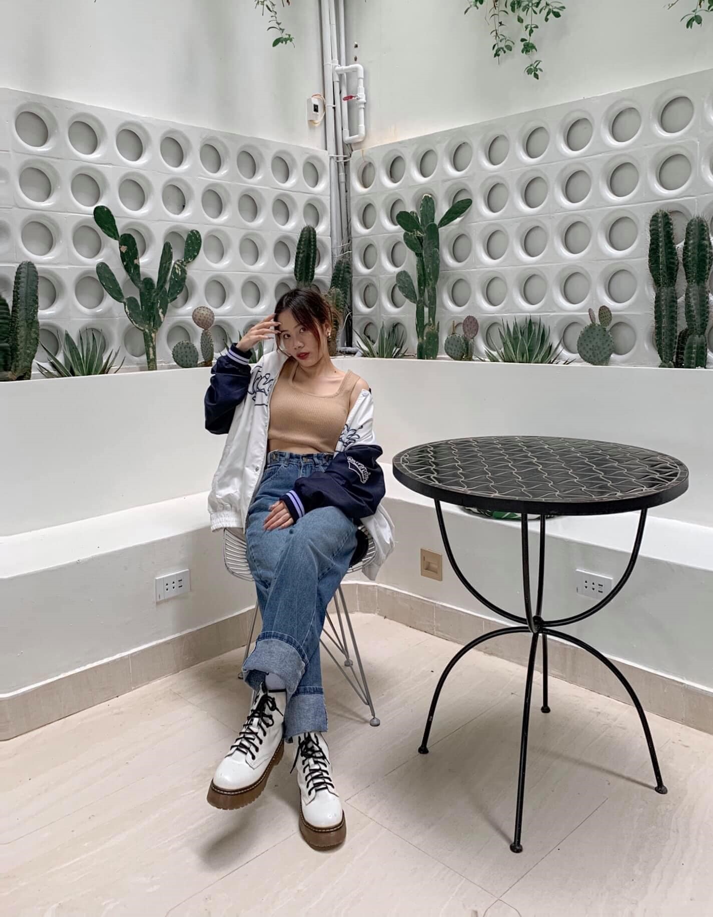 Người ngồi tr&ecirc;n ghế cạnh b&agrave;n tr&ograve;n, nền l&agrave; tường trang tr&iacute; với c&acirc;y xương rồng v&agrave; hoạ tiết tr&ograve;n.