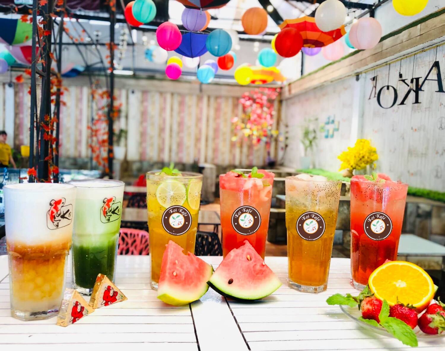 C&aacute;c ly nước tr&aacute;i c&acirc;y đa dạng k&egrave;m l&aacute;t dưa hấu v&agrave; cam, b&agrave;y tr&ecirc;n b&agrave;n trong kh&ocirc;ng gian trang tr&iacute; đ&egrave;n lồng sặc sỡ.