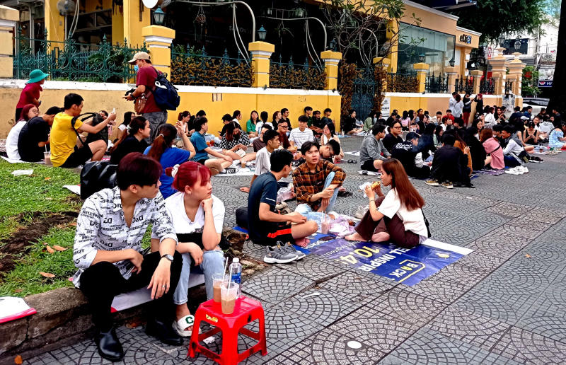 Một nh&oacute;m đ&ocirc;ng người ngồi tr&ecirc;n vỉa h&egrave;, tr&ograve; chuyện v&agrave; ăn uống trước một t&ograve;a nh&agrave; m&agrave;u v&agrave;ng.