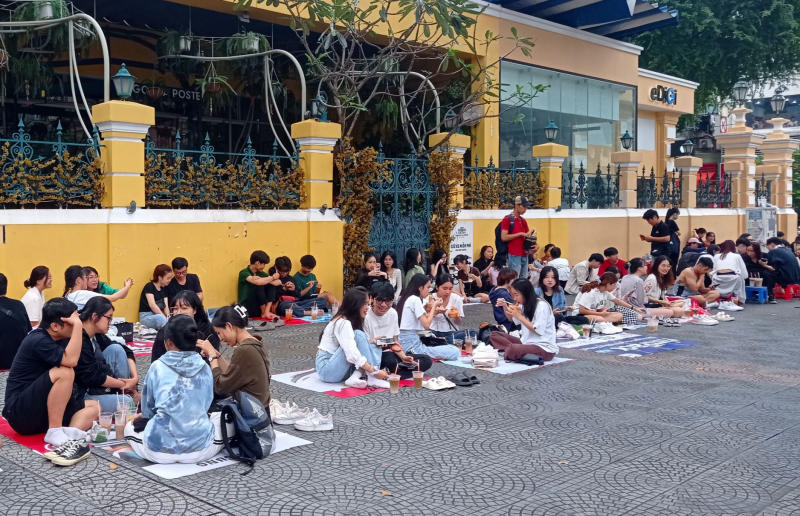 Một nh&oacute;m người ngồi tr&ecirc;n vỉa h&egrave; trước một t&ograve;a nh&agrave; m&agrave;u v&agrave;ng, thưởng thức đồ ăn v&agrave; thức uống.