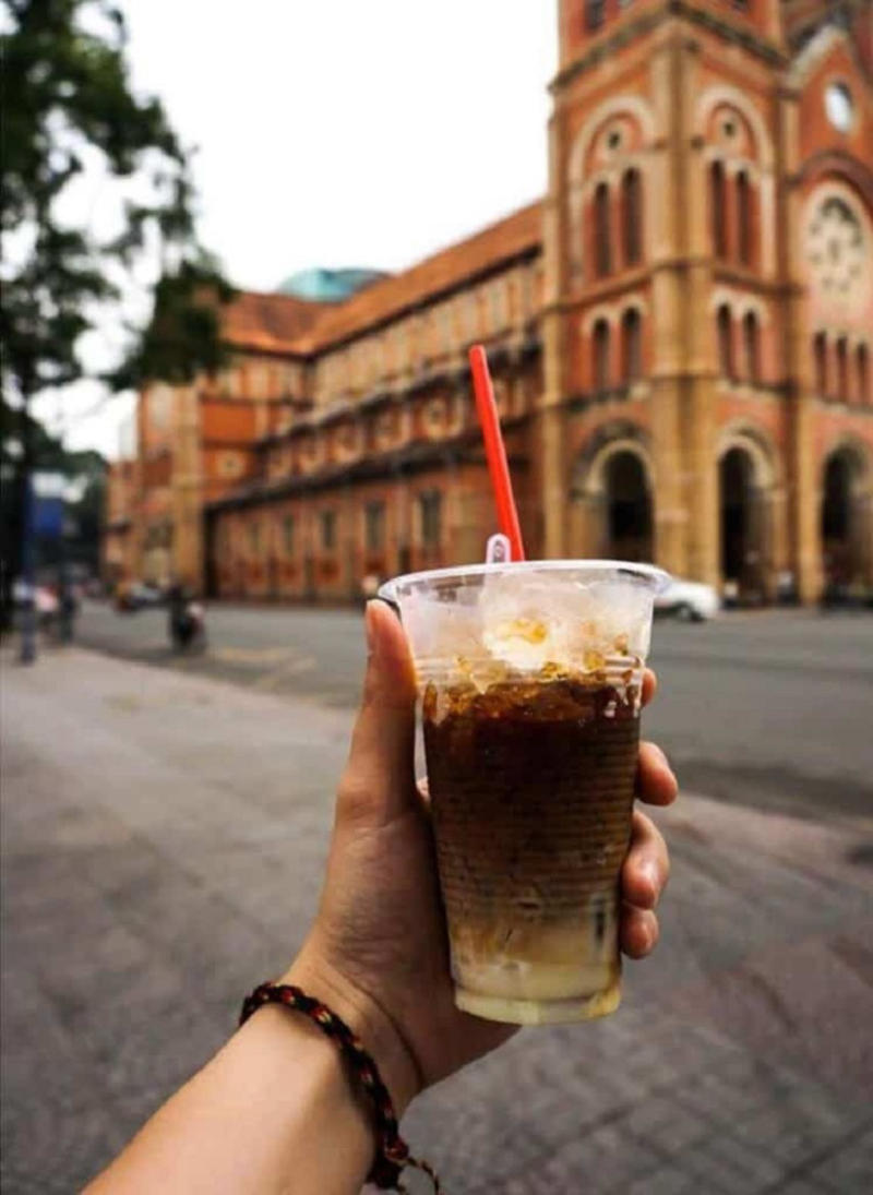 Tay cầm ly c&agrave; ph&ecirc; sữa đ&aacute; trước một t&ograve;a nh&agrave; cổ k&iacute;nh bằng gạch đỏ ở khu đ&ocirc; thị.