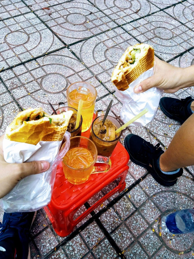 Hai người cầm b&aacute;nh m&igrave;, b&ecirc;n cạnh l&agrave; b&agrave;n nhựa đỏ với ba ly nước tr&ecirc;n vỉa h&egrave; l&aacute;t gạch.