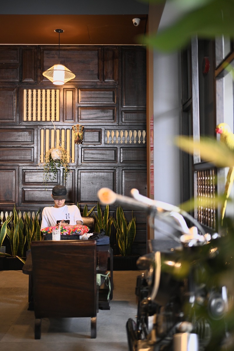 Một người ngồi tr&ecirc;n ghế gỗ, sử dụng điện thoại, ph&iacute;a sau l&agrave; tường gỗ trang tr&iacute; v&agrave; c&acirc;y xanh.