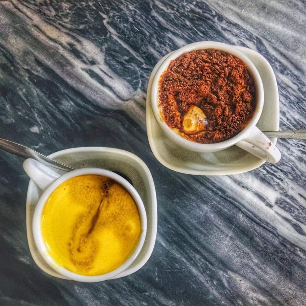 Hai t&aacute;ch c&agrave; ph&ecirc; trứng tr&ecirc;n b&agrave;n đ&aacute; cẩm thạch, một t&aacute;ch phủ bột cacao v&agrave; một t&aacute;ch c&oacute; bề mặt v&agrave;ng mịn.