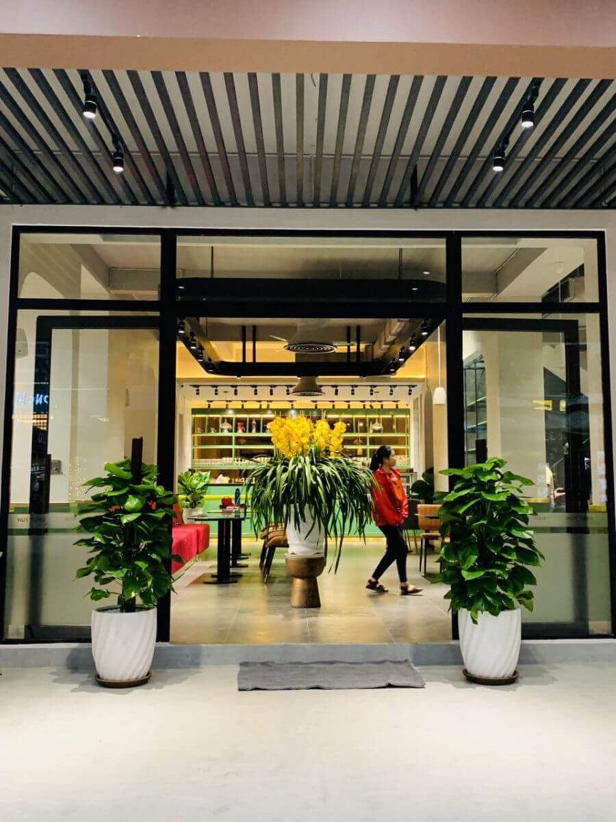 Lối v&agrave;o một qu&aacute;n c&agrave; ph&ecirc; với c&acirc;y xanh v&agrave; hoa trang tr&iacute;, c&oacute; người đi bộ b&ecirc;n trong.