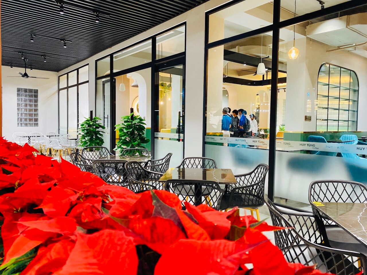 Qu&aacute;n c&agrave; ph&ecirc; với b&agrave;n ghế ngo&agrave;i trời, c&acirc;y xanh v&agrave; hoa đỏ trang tr&iacute;, nh&igrave;n v&agrave;o b&ecirc;n trong qua cửa k&iacute;nh.