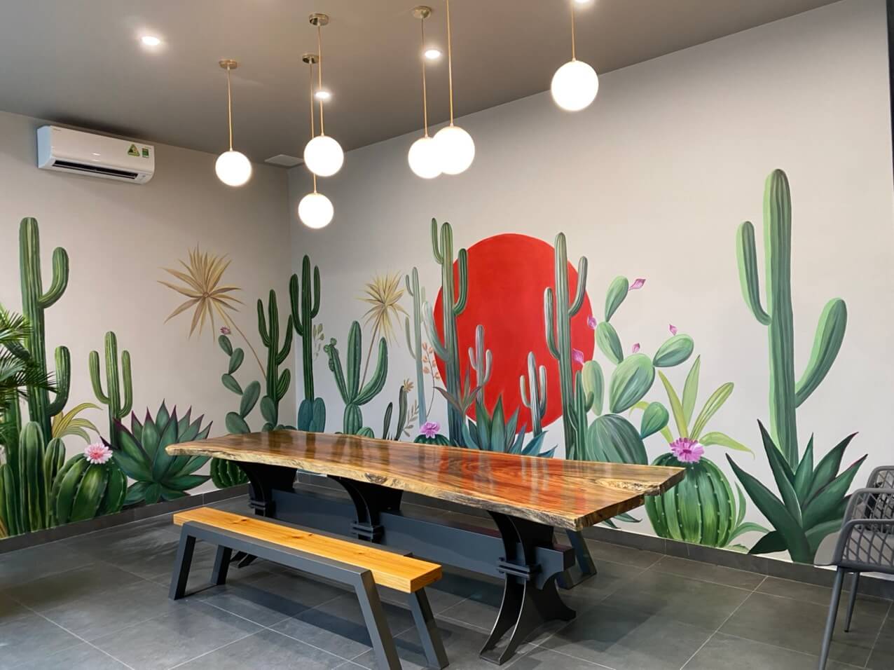 Ph&ograve;ng c&oacute; bức tường vẽ xương rồng v&agrave; mặt trời đỏ, b&agrave;n gỗ d&agrave;i với ghế, đ&egrave;n treo trần v&agrave; m&aacute;y lạnh.