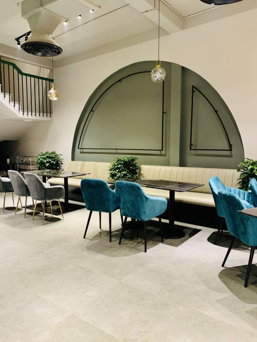 Qu&aacute;n c&agrave; ph&ecirc; hiện đại với b&agrave;n ghế m&agrave;u x&aacute;m v&agrave; xanh, c&acirc;y trang tr&iacute;, &aacute;nh s&aacute;ng đ&egrave;n treo v&agrave; cửa sổ h&igrave;nh v&ograve;m.