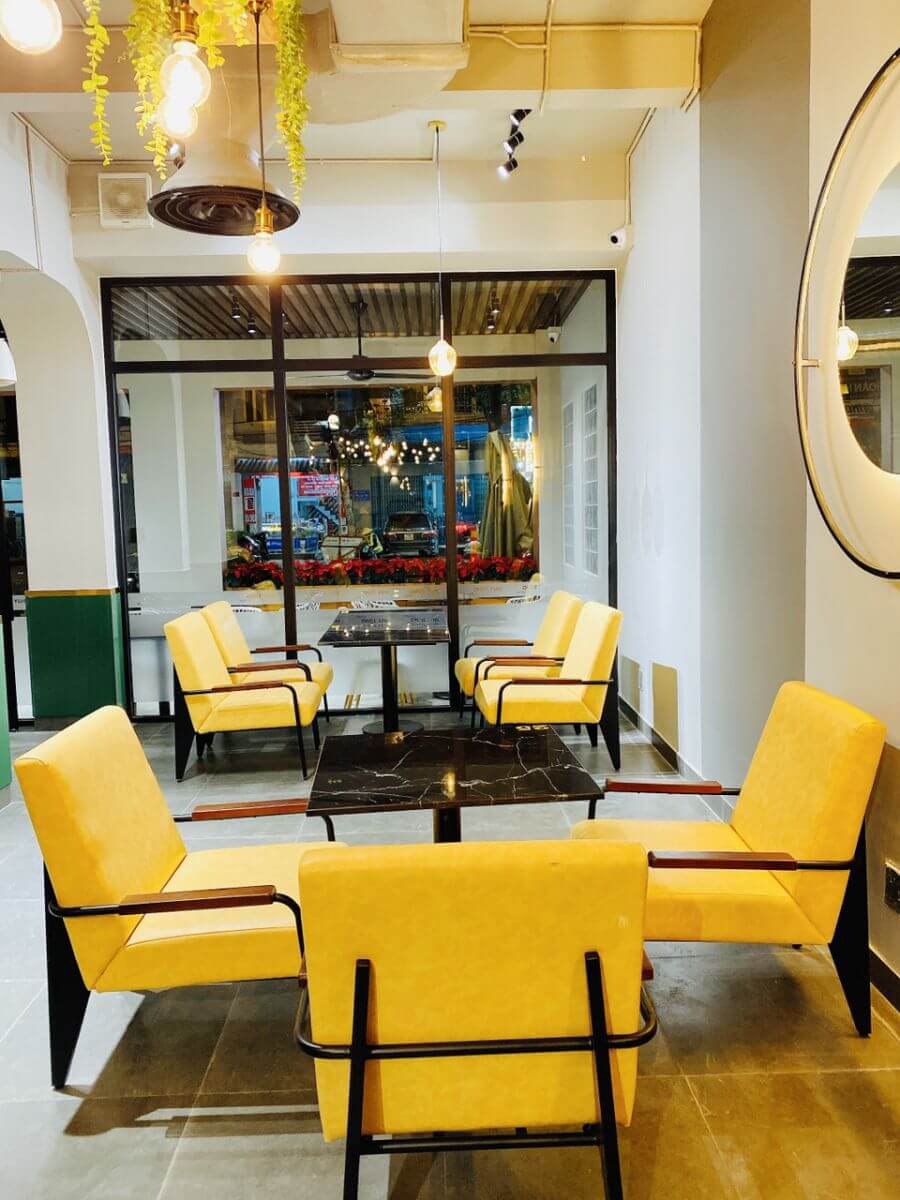 Nội thất qu&aacute;n c&agrave; ph&ecirc; với ghế m&agrave;u v&agrave;ng tươi, b&agrave;n đen, &aacute;nh s&aacute;ng ấm &aacute;p v&agrave; trang tr&iacute; c&acirc;y xanh.
