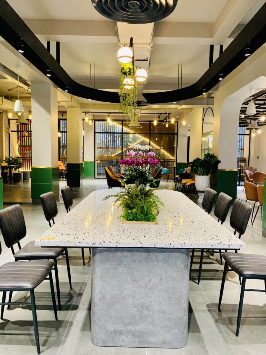 Kh&ocirc;ng gian nội thất hiện đại với b&agrave;n d&agrave;i, ghế ngồi v&agrave; c&acirc;y xanh trang tr&iacute; dưới &aacute;nh s&aacute;ng đ&egrave;n trần.