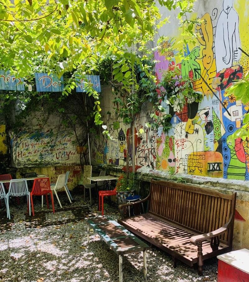 Không gian ngoài trời với ghế gỗ, ghế nhựa và tường vẽ graffiti, cây xanh che bóng mát.