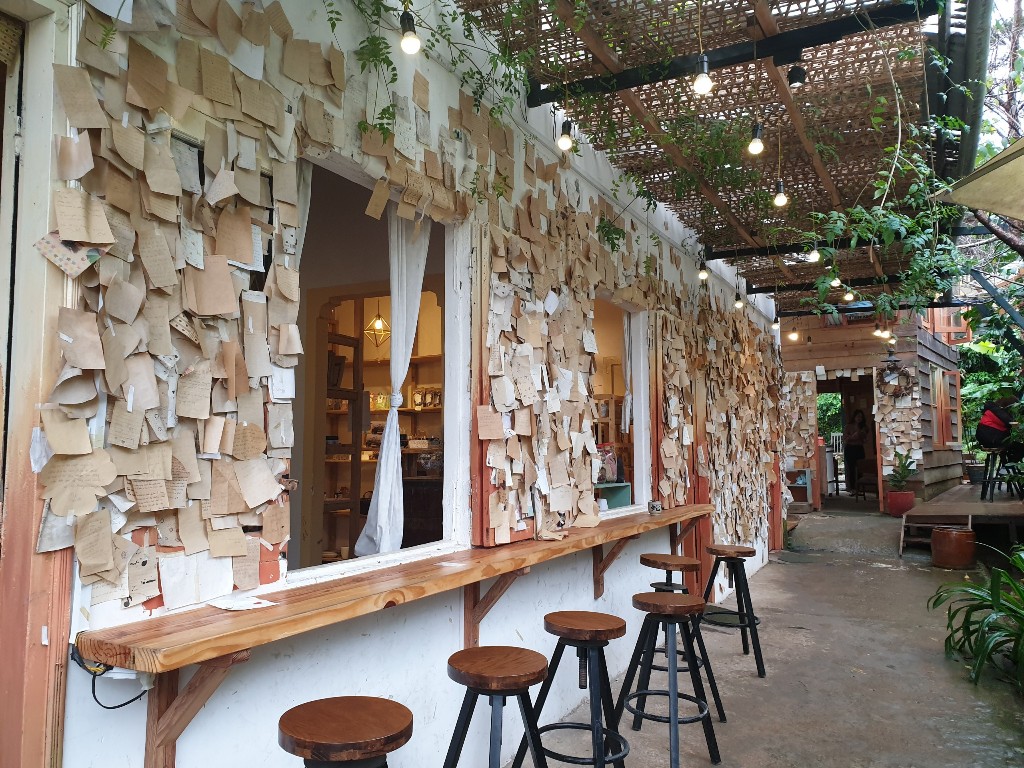 Qu&aacute;n c&agrave; ph&ecirc; với tường d&aacute;n đầy giấy ghi ch&uacute;, b&agrave;n gỗ v&agrave; ghế cao, &aacute;nh s&aacute;ng đ&egrave;n treo v&agrave; c&acirc;y xanh trang tr&iacute;.