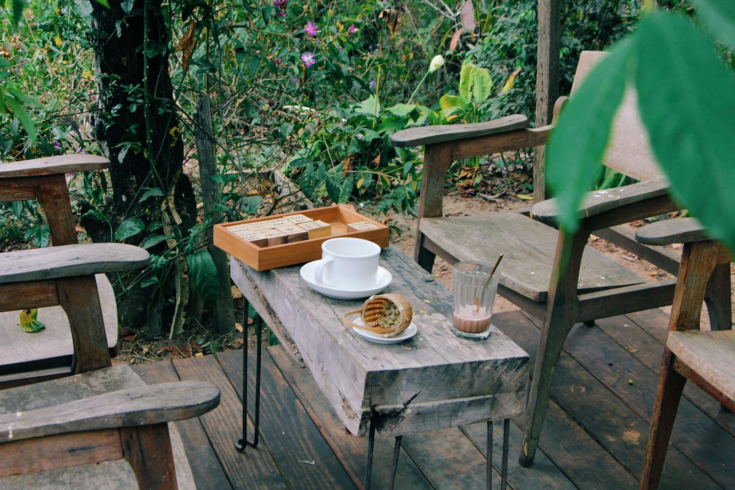 B&agrave;n gỗ ngo&agrave;i trời với t&aacute;ch tr&agrave;, ly c&agrave; ph&ecirc; v&agrave; bộ tr&ograve; chơi, xung quanh l&agrave; ghế gỗ trong kh&ocirc;ng gian xanh.