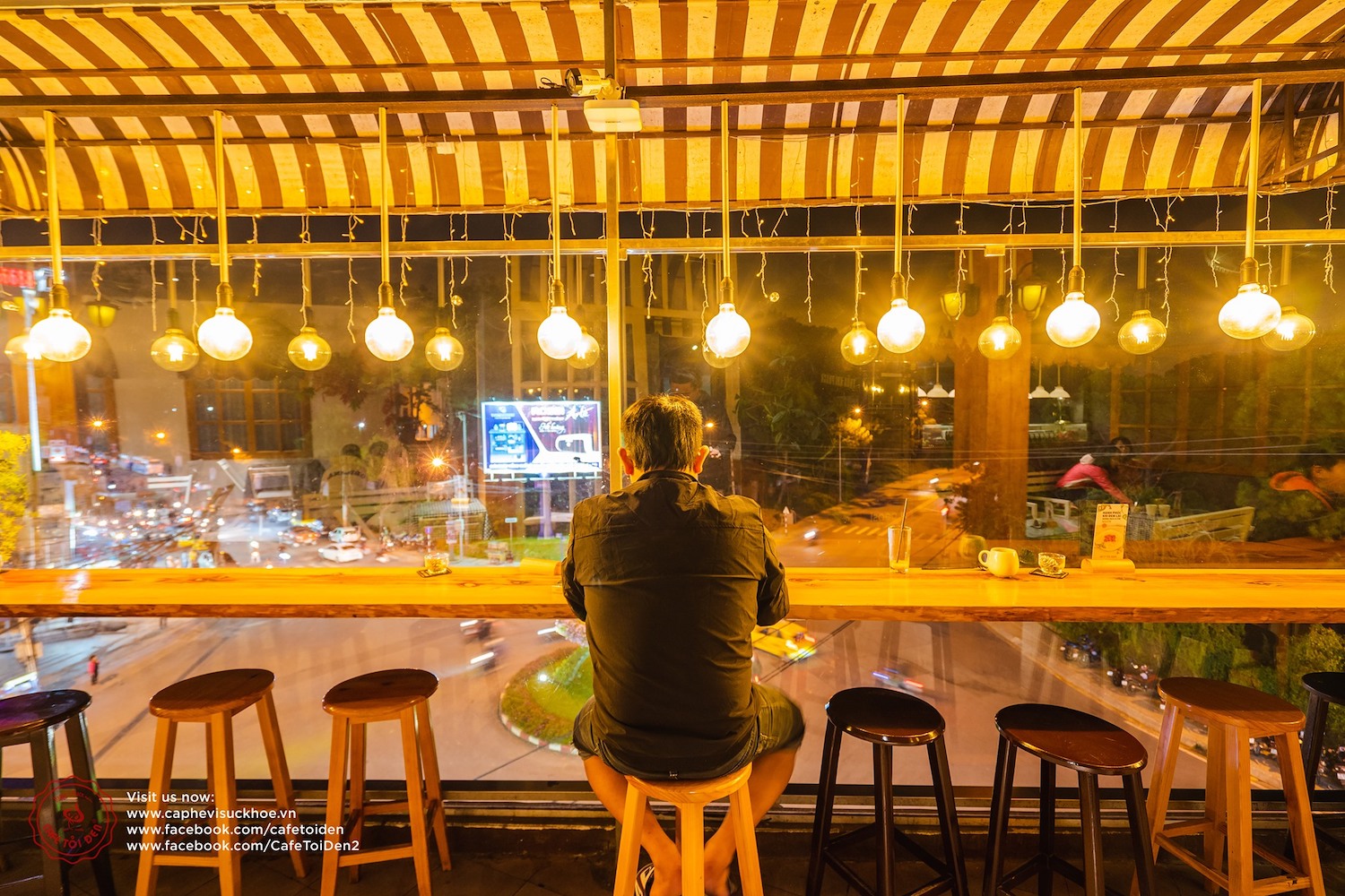 Người ngồi tại quán cà phê có cửa kính lớn nhìn ra đường phố sáng đèn vào ban đêm, bên trong có đèn trang trí.