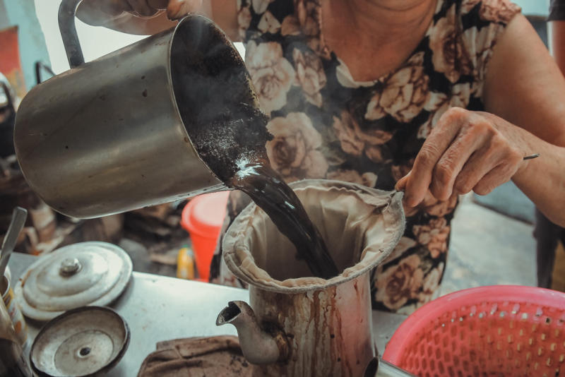 Người đang rót cà phê đen từ ấm kim loại qua túi lọc vào bình, trên bàn có nắp nồi.