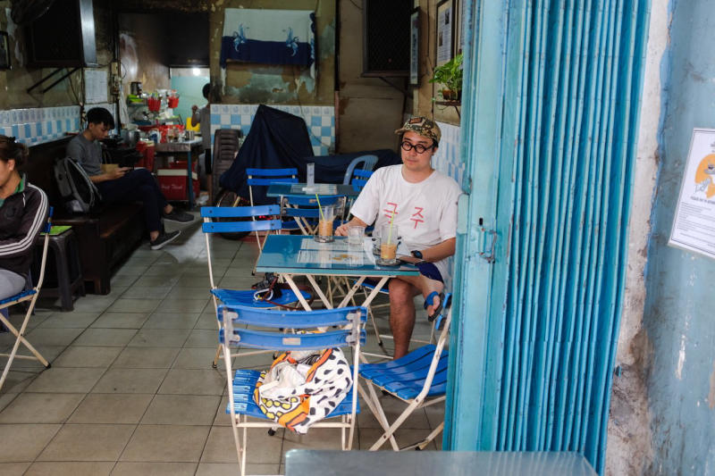 Quán cà phê nhỏ với bàn ghế xanh, có người ngồi uống nước. Nội thất đơn giản và không gian ấm cúng.