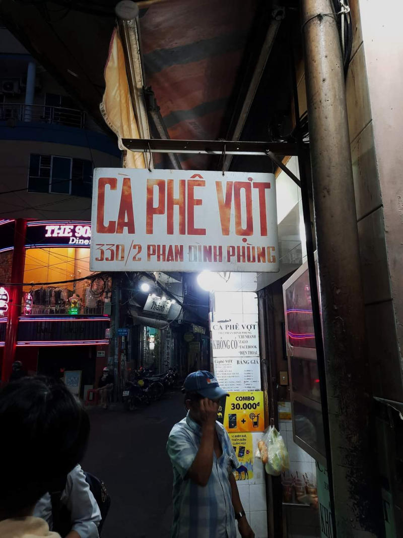 Biển hiệu "Cà Phê Vợt" treo trước một quán cà phê nhỏ trong khu phố, có người đang đứng gần đó.