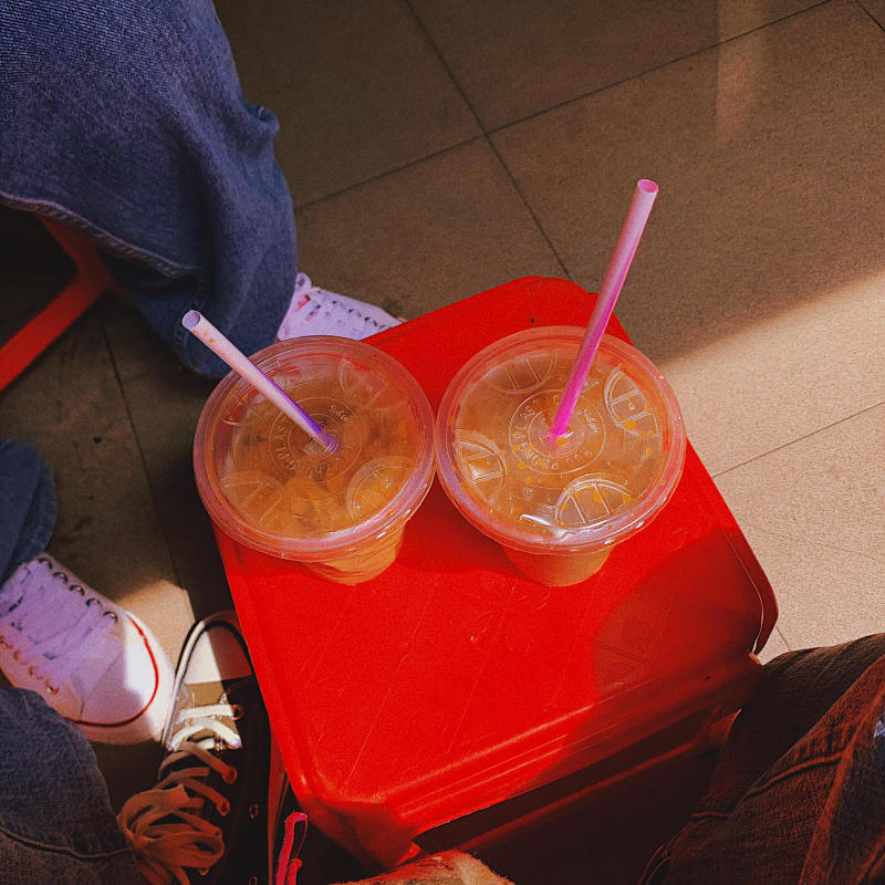 Hai cốc nước có ống hút trên ghế nhựa đỏ, cạnh đôi giày thể thao trên nền gạch.