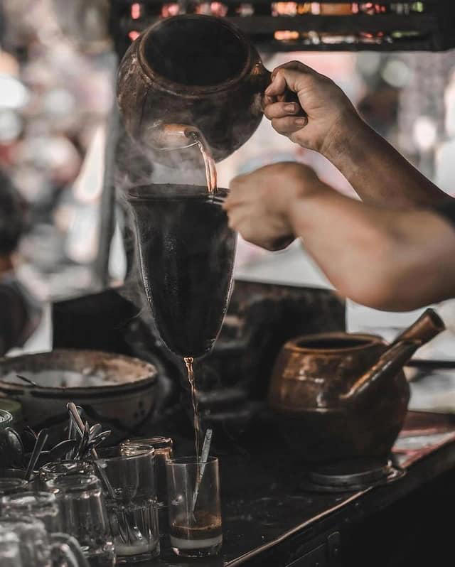 Người đang rót nước từ ấm đất qua một bộ lọc vải vào ly thủy tinh, xung quanh là nhiều ly và bình khác.