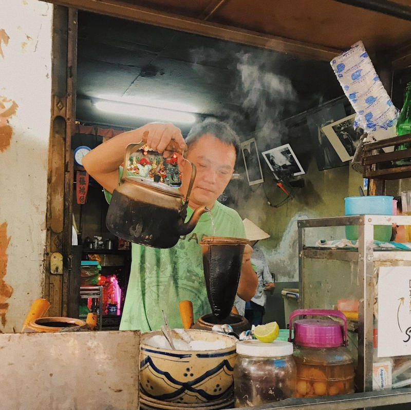 Người đàn ông đang pha chế cà phê bằng ấm đun nước tại một quán nhỏ, xung quanh có nhiều vật dụng pha chế.