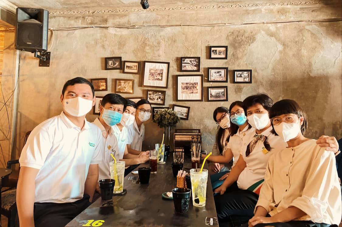 Nh&oacute;m người đeo khẩu trang ngồi quanh b&agrave;n trong qu&aacute;n c&agrave; ph&ecirc;, tr&ecirc;n tường c&oacute; nhiều bức ảnh trang tr&iacute;.