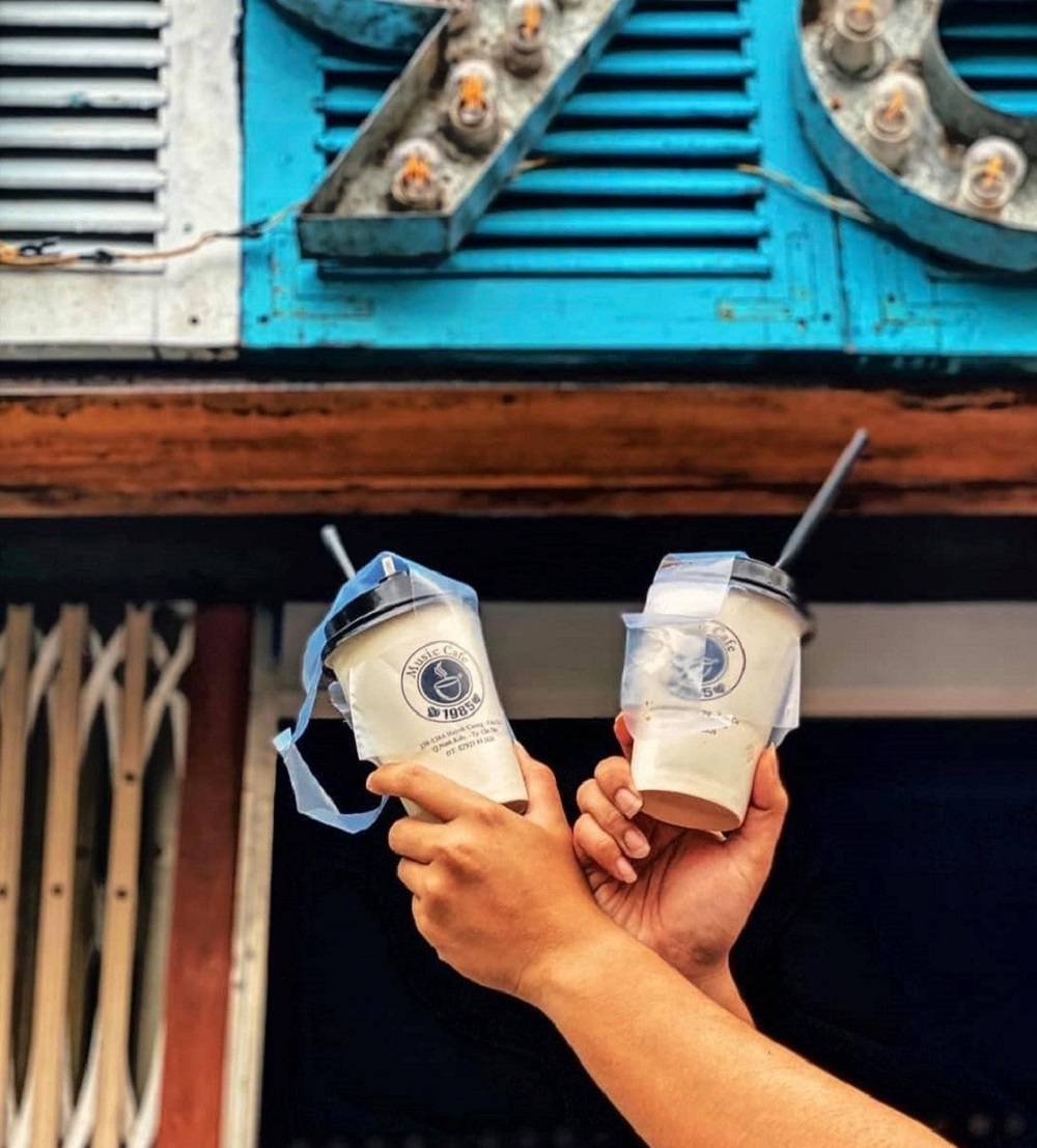 Hai tay giơ cao hai ly nhựa c&oacute; ống h&uacute;t trước một biển hiệu m&agrave;u xanh lam với đ&egrave;n tr&ograve;n.