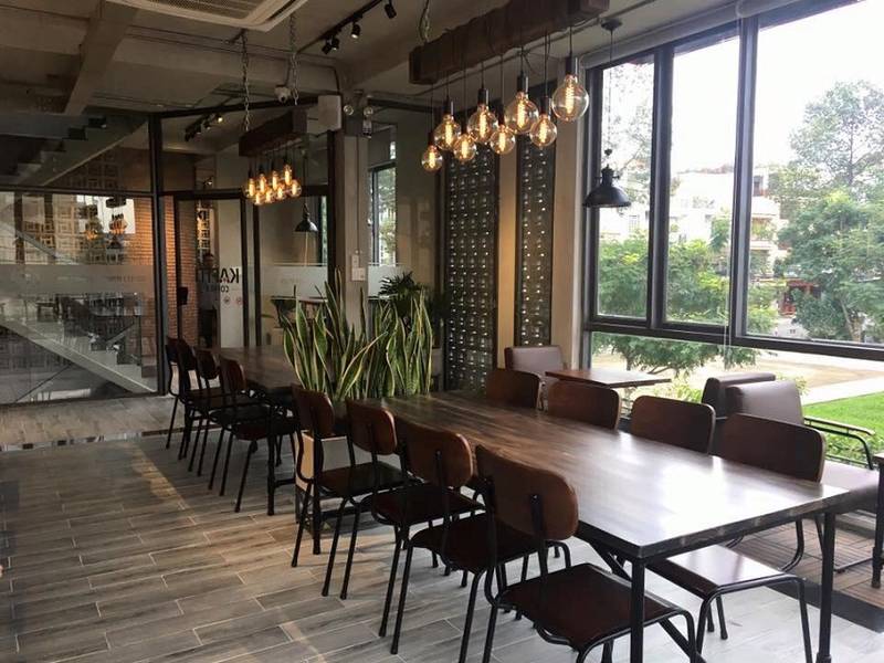 Qu&aacute;n c&agrave; ph&ecirc; với b&agrave;n gỗ d&agrave;i, ghế xếp gọn g&agrave;ng, đ&egrave;n treo trang tr&iacute; v&agrave; c&acirc;y xanh, cửa sổ lớn nh&igrave;n ra b&ecirc;n ngo&agrave;i.