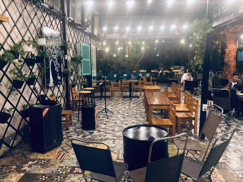 Qu&aacute;n c&agrave; ph&ecirc; ngo&agrave;i trời với b&agrave;n ghế gỗ, trang tr&iacute; c&acirc;y xanh v&agrave; &aacute;nh đ&egrave;n, c&oacute; v&agrave;i người ngồi tr&ograve; chuyện.