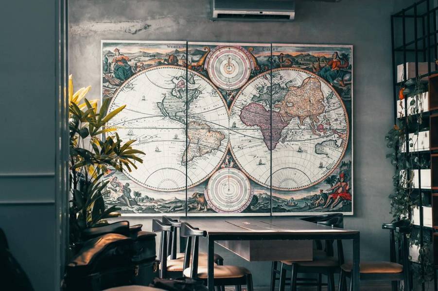 Bức tranh bản đồ cổ treo tr&ecirc;n tường trong một qu&aacute;n c&agrave; ph&ecirc;, b&ecirc;n cạnh l&agrave; b&agrave;n ghế v&agrave; c&acirc;y xanh trang tr&iacute;.