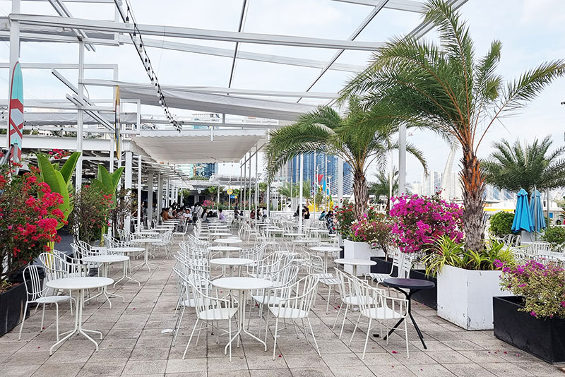 Kh&ocirc;ng gian ngo&agrave;i trời với b&agrave;n ghế trắng, c&acirc;y cọ v&agrave; hoa trang tr&iacute;, tạo cảm gi&aacute;c thư gi&atilde;n v&agrave; tho&aacute;ng đ&atilde;ng.