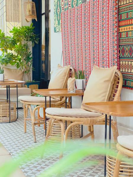 Qu&aacute;n c&agrave; ph&ecirc; với b&agrave;n ghế m&acirc;y, đệm m&agrave;u be, nền gạch hoa v&agrave; tường treo vải trang tr&iacute; họa tiết m&agrave;u sắc.