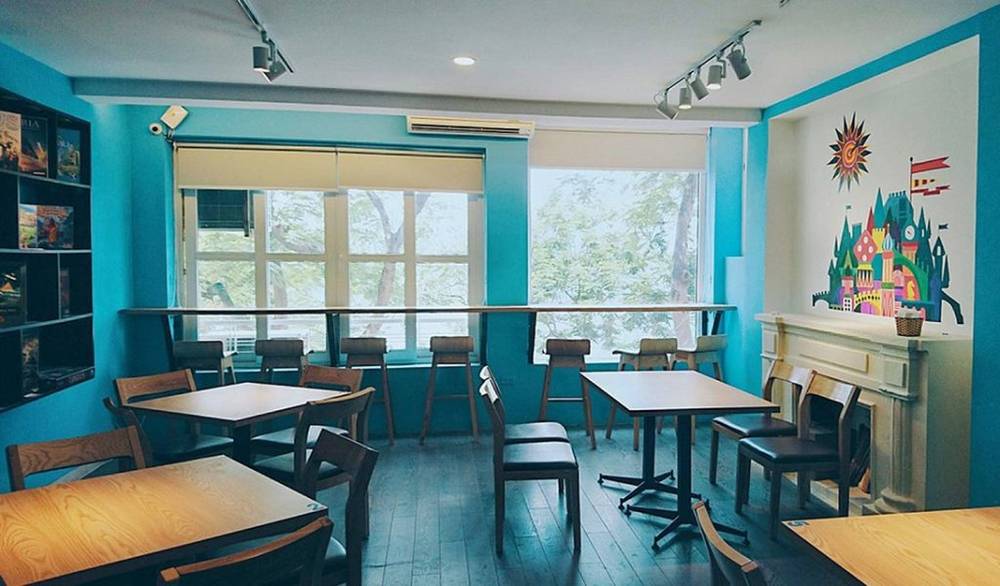 Ph&ograve;ng c&agrave; ph&ecirc; với tường xanh, b&agrave;n ghế gỗ, cửa sổ lớn, v&agrave; bức tranh tường nhiều m&agrave;u sắc.