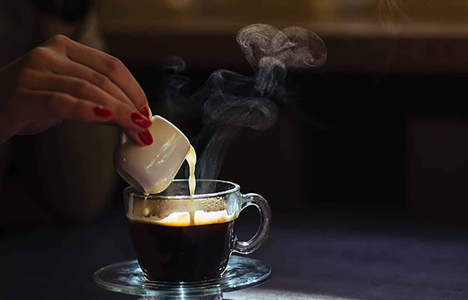 Tay đổ sữa v&agrave;o t&aacute;ch c&agrave; ph&ecirc; n&oacute;ng bốc kh&oacute;i, tr&ecirc;n b&agrave;n c&oacute; &aacute;nh s&aacute;ng nhẹ nh&agrave;ng chiếu v&agrave;o.