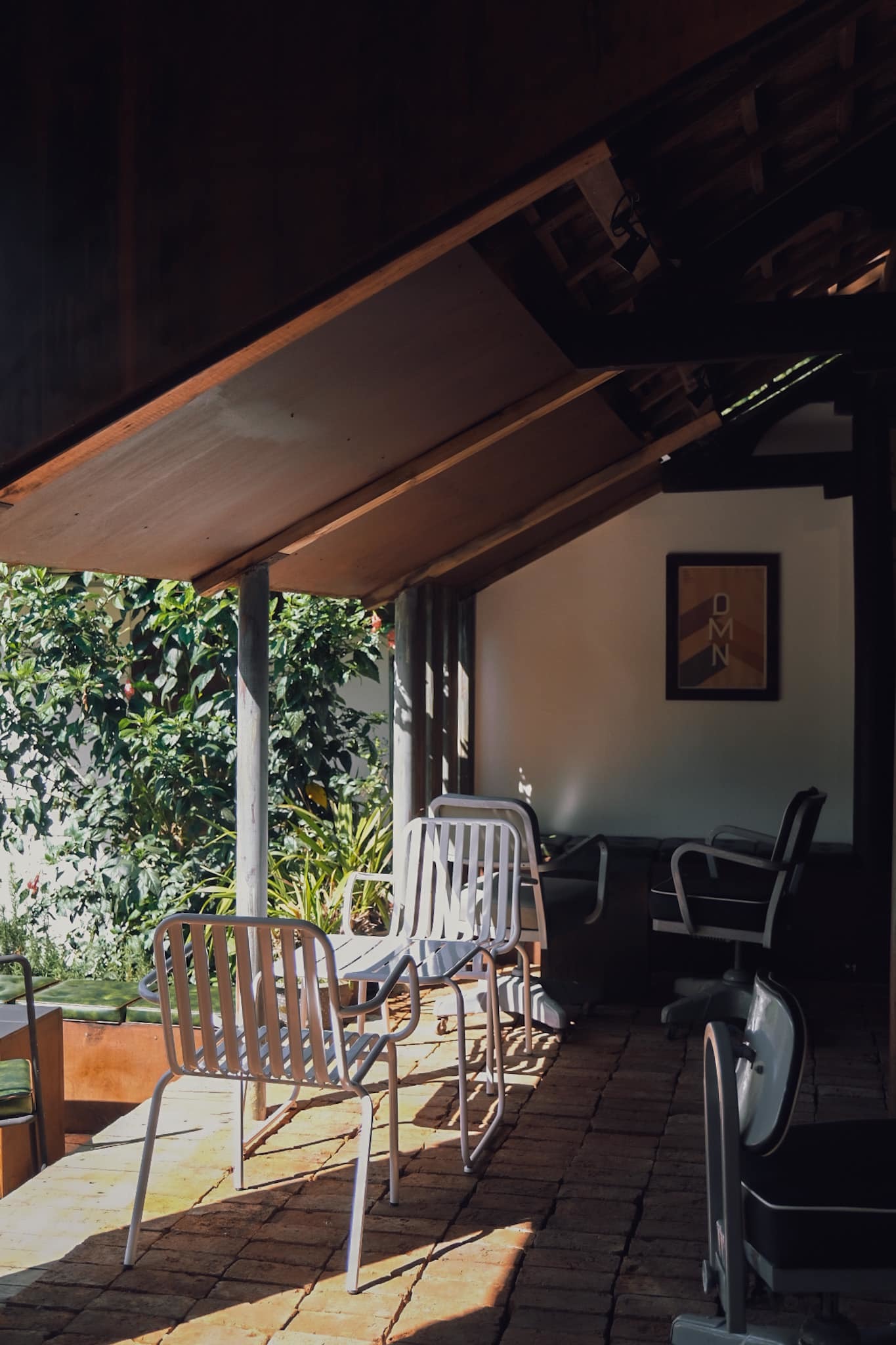 Ghế kim loại v&agrave; ghế xoay đặt dưới m&aacute;i hi&ecirc;n gỗ, gần tường c&oacute; c&acirc;y xanh v&agrave; tranh treo.