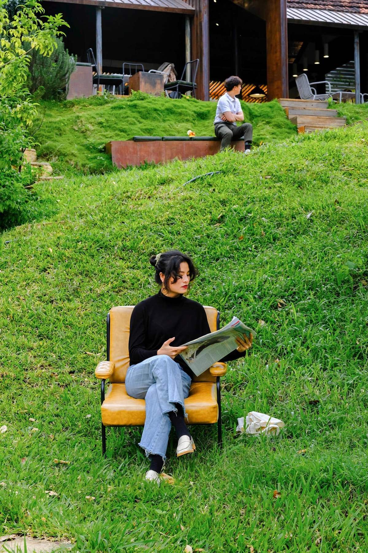 Người ngồi tr&ecirc;n ghế v&agrave;ng đọc b&aacute;o, ph&iacute;a sau l&agrave; một người kh&aacute;c ngồi tr&ecirc;n bậc thềm, cảnh quan s&acirc;n cỏ xanh.