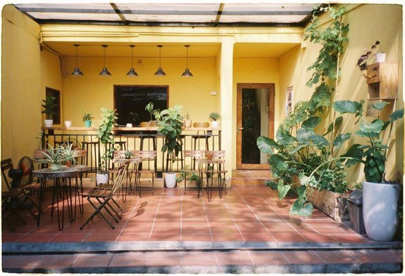 Qu&aacute;n c&agrave; ph&ecirc; ngo&agrave;i trời với tường v&agrave;ng, b&agrave;n ghế kim loại, c&acirc;y xanh trang tr&iacute; v&agrave; &aacute;nh s&aacute;ng tự nhi&ecirc;n.