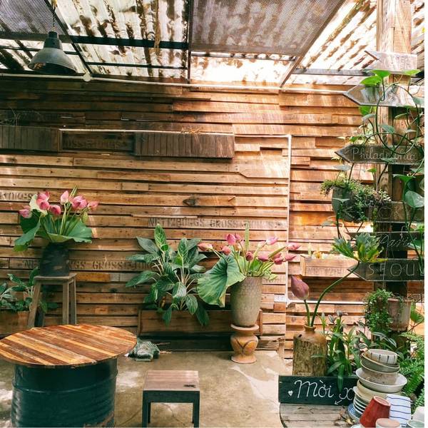 Kh&ocirc;ng gian qu&aacute;n c&agrave; ph&ecirc; với tường gỗ, b&agrave;n tr&ograve;n, chậu c&acirc;y xanh v&agrave; hoa trang tr&iacute;, tạo cảm gi&aacute;c mộc mạc, gần gũi.