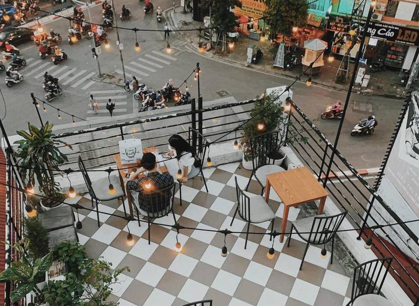 Qu&aacute;n c&agrave; ph&ecirc; tr&ecirc;n cao với b&agrave;n ghế ngo&agrave;i trời, đ&egrave;n treo lấp l&aacute;nh, nh&igrave;n xuống đường phố nhộn nhịp xe cộ.