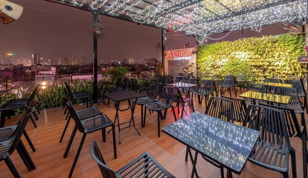 Qu&aacute;n c&agrave; ph&ecirc; s&acirc;n thượng với b&agrave;n ghế đen, đ&egrave;n trang tr&iacute; lấp l&aacute;nh v&agrave; bức tường c&acirc;y xanh, nh&igrave;n ra th&agrave;nh phố ban đ&ecirc;m.