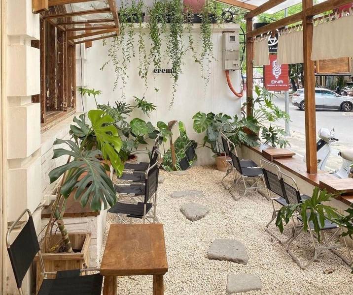 Qu&aacute;n c&agrave; ph&ecirc; ngo&agrave;i trời với b&agrave;n ghế gỗ, c&acirc;y xanh trang tr&iacute;, sỏi trắng trải nền, kh&ocirc;ng gian tho&aacute;ng đ&atilde;ng b&ecirc;n lề đường.