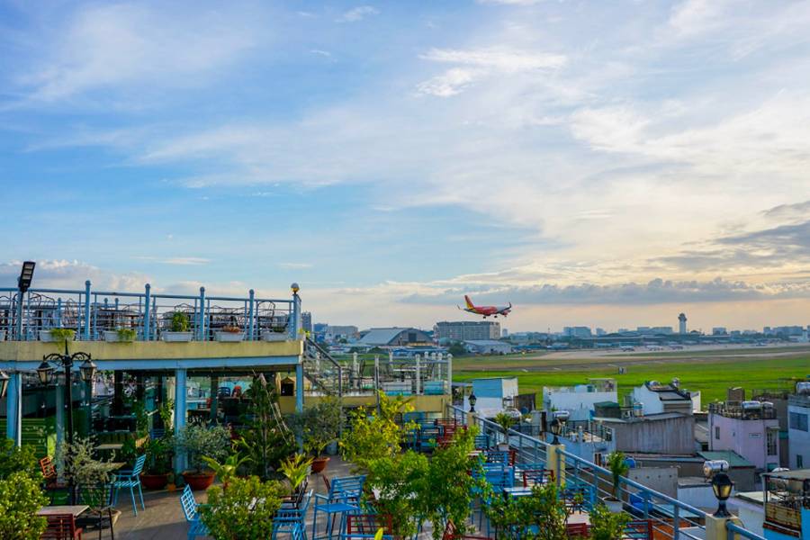 Quán cà phê sân thượng với bàn ghế nhiều màu sắc, máy bay đang hạ cánh trên sân bay gần đó, trời xanh mây trắng.