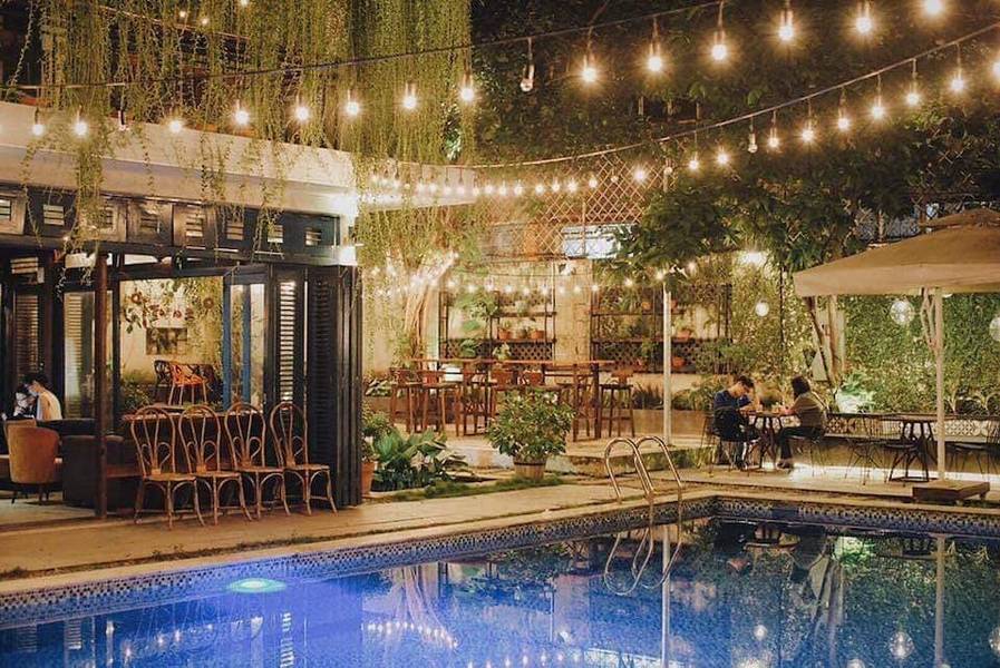 Quán cà phê ngoài trời với hồ bơi, trang trí đèn lấp lánh và cây xanh, khách đang ngồi trò chuyện.