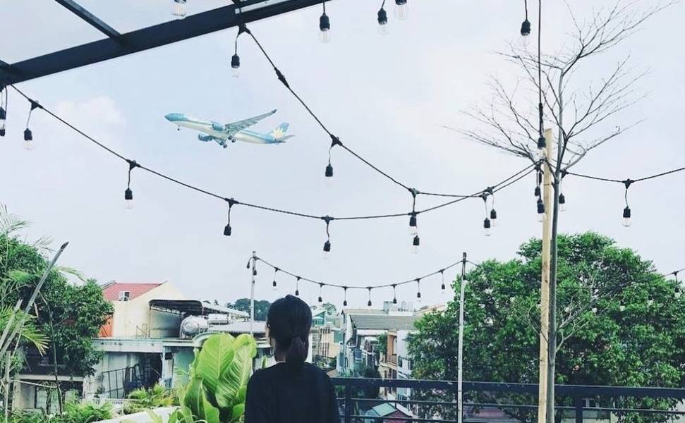 Người đứng trên ban công nhìn máy bay bay qua, xung quanh là dây đèn trang trí và cây xanh.
