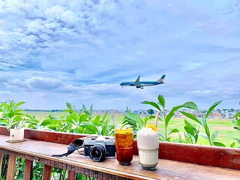 Máy bay bay trên bầu trời xanh, phía dưới là hai ly nước và một máy ảnh trên bàn gỗ gần cây xanh.