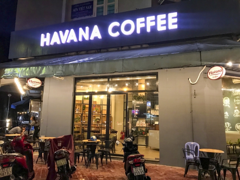 Mặt tiền quán cà phê với bảng hiệu sáng đèn, bàn ghế ngoài trời và xe máy đậu trước cửa vào ban đêm.