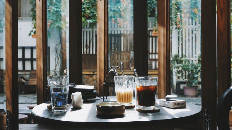 Bàn cà phê với ba ly nước và gạt tàn, ánh sáng tự nhiên chiếu qua cửa kính, khung cảnh ngoài trời có cây xanh.